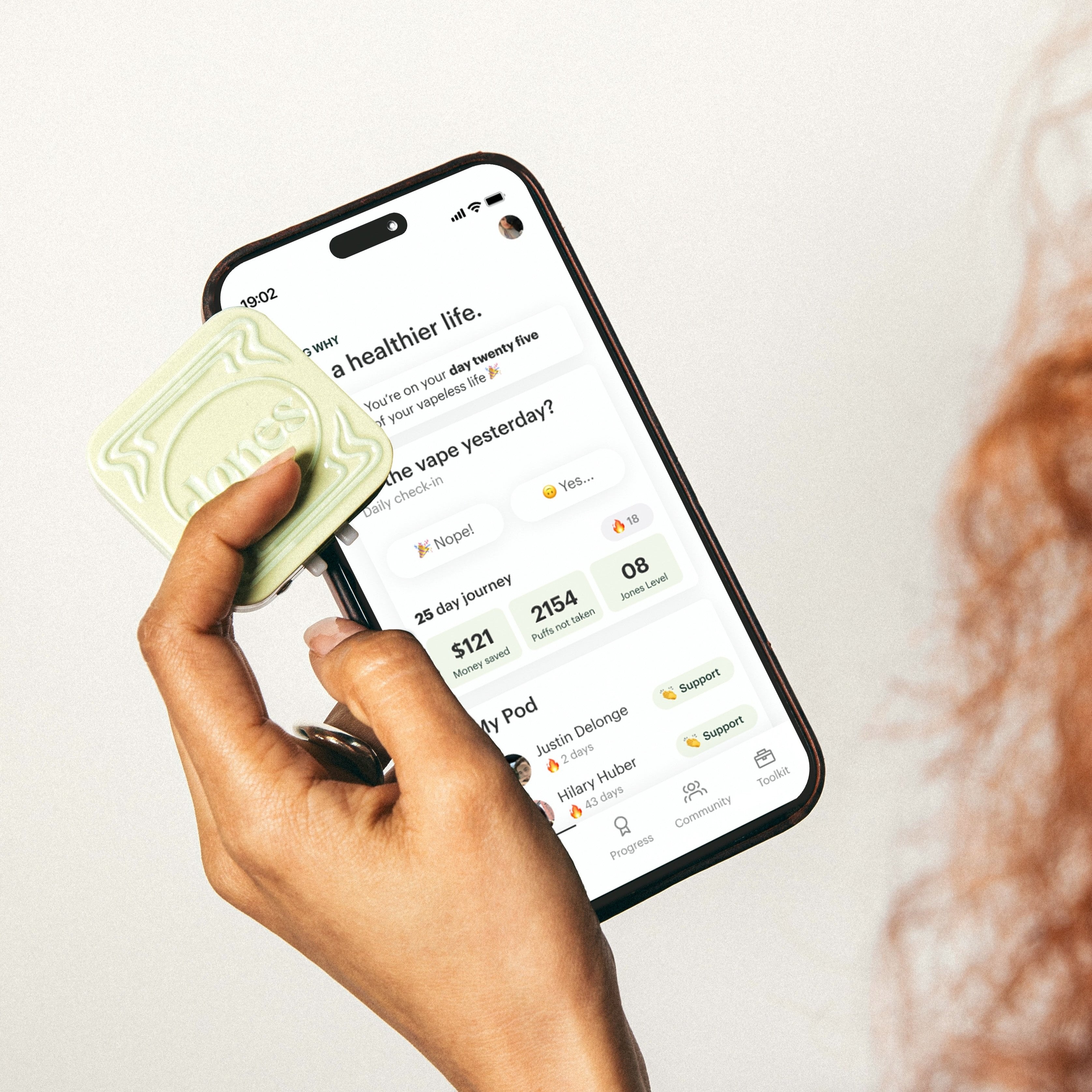 A hand holding a small square metal tin with embellishments labeled Jones that contains FDA-approved Jones nicotine replacement therapy lozenges and a cell phone screen showing an app to quit vaping by Jones with money saved, streak, and daily nicotine tracking.