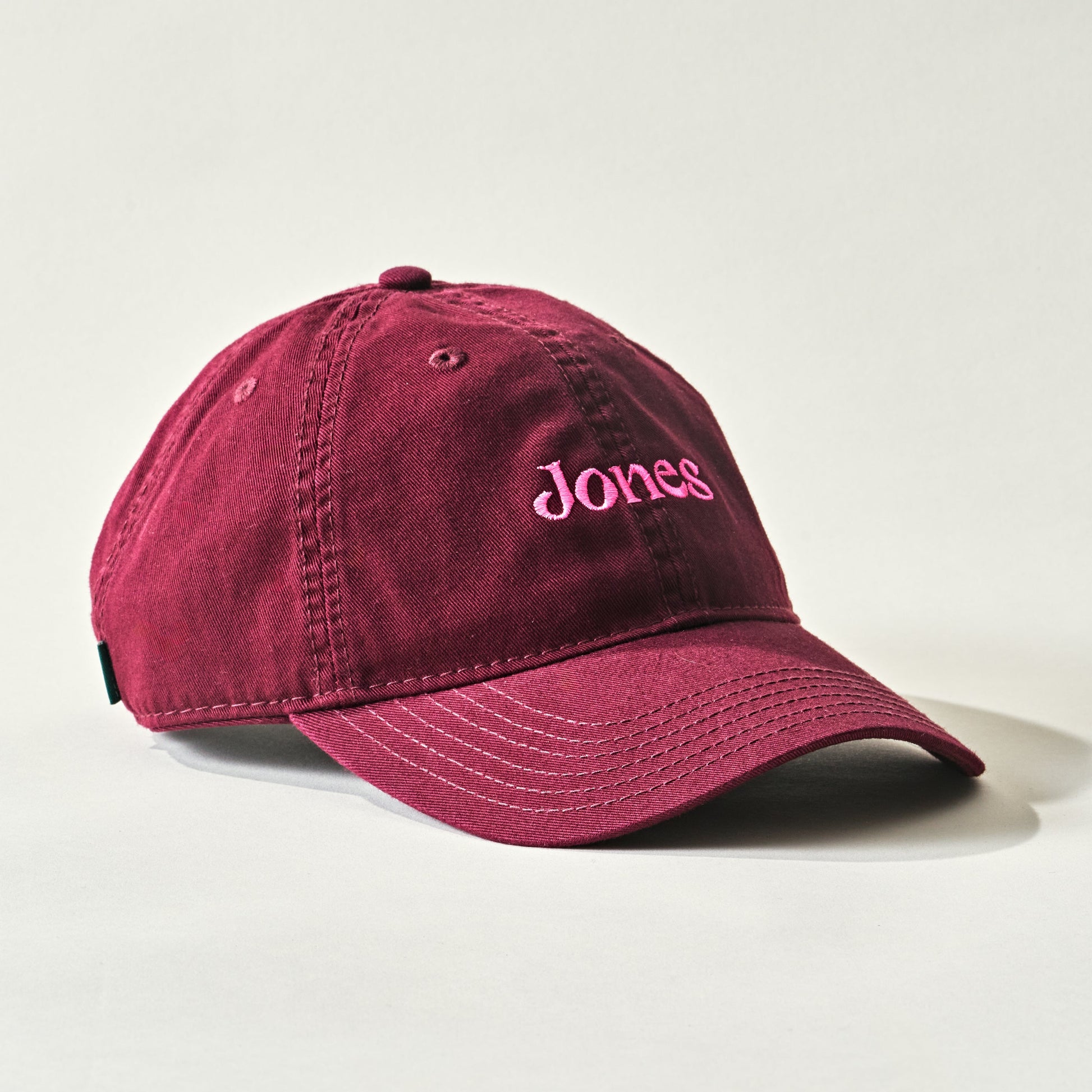 The Jones Cap by Quit with Jones is a maroon cotton twill baseball cap featuring "Jones" embroidered in pink on the front, shown on a light background.