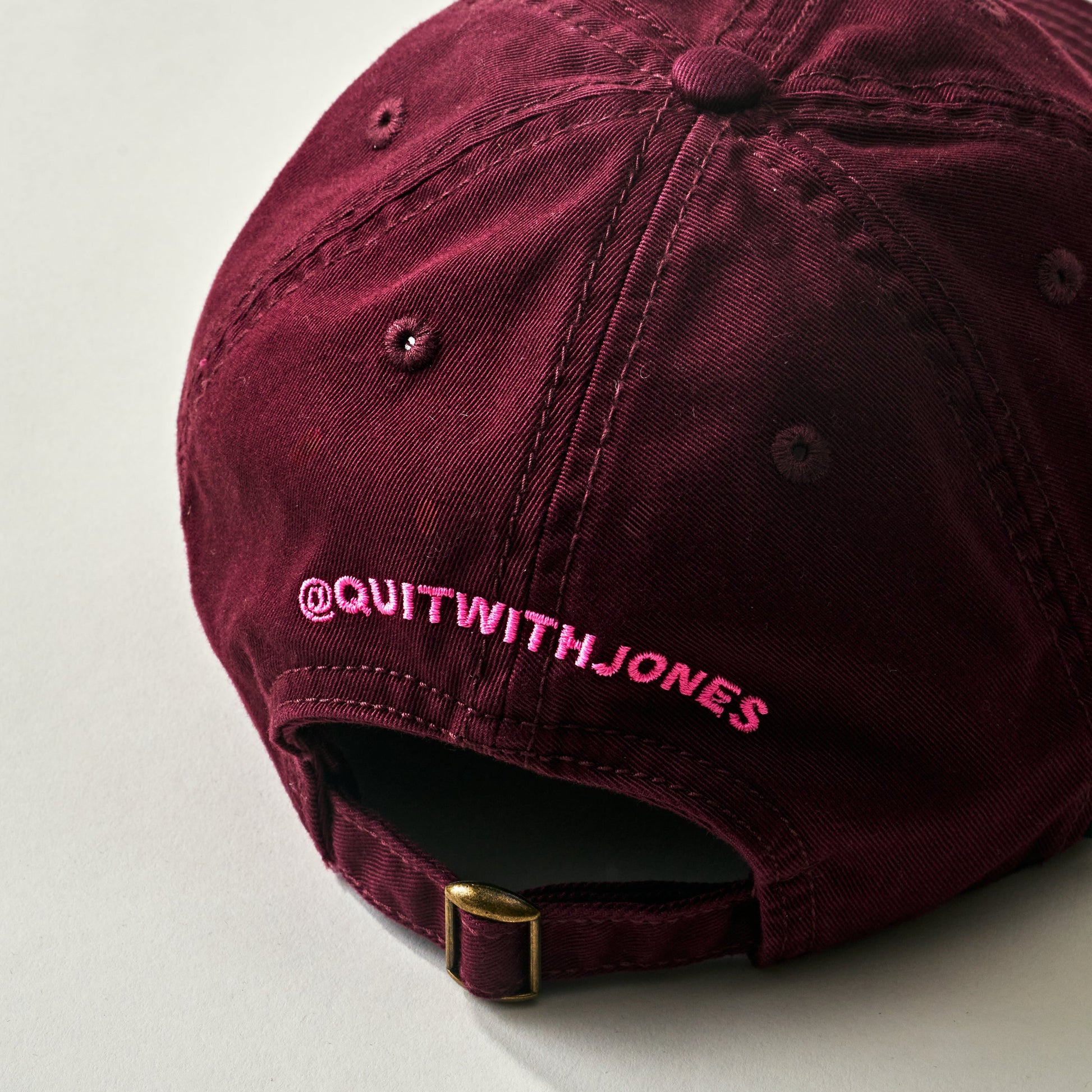 The Jones Cap by Quit with Jones is a maroon cotton twill baseball cap featuring "Jones" embroidered in pink on the front, shown on a light background.