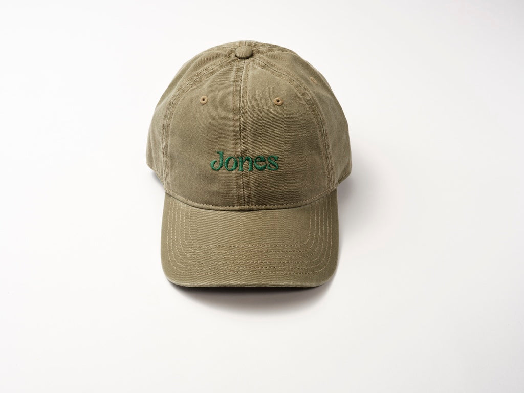 A khaki cotton twill baseball cap, named the Jones Cap by Quit with Jones, features "Jones" embroidered in moss green on the front and rests on a plain white surface.