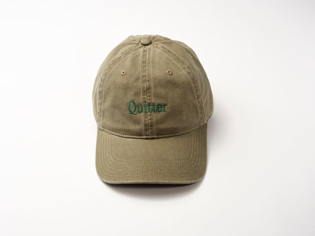 The Quitter Cap by Quit with Jones is a khaki cotton twill baseball cap featuring "Quitter" embroidered in moss green on the front, set against a plain white background.