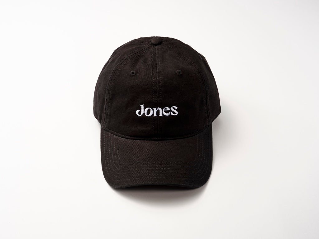 The Jones Cap by Quit with Jones is a black cotton twill baseball hat featuring "Jones" embroidered in white on the front, displayed on a white surface.
