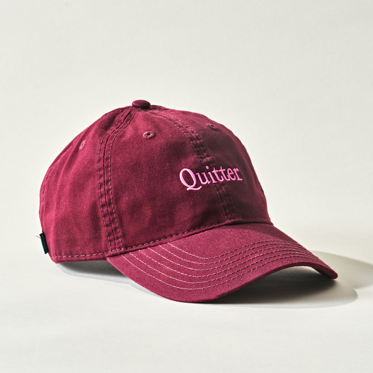 The Quitter Cap by Quit with Jones is a maroon cotton twill baseball cap featuring “Quitter” embroidered in pink on the front, shown against a plain light background.
