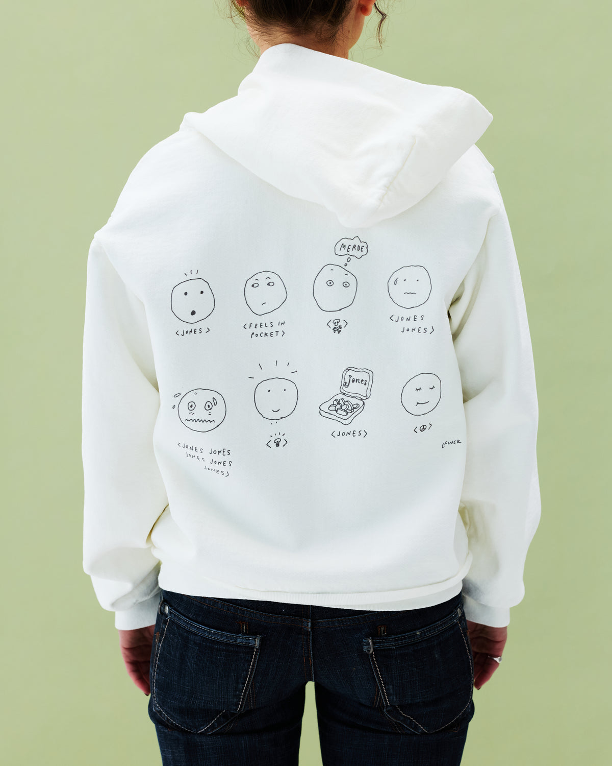 A person in blue jeans faces away, wearing the Jones / Liana Finck Jonesing Hoodie by Quit with Jones. The unisex premium cotton hoodie features nine cartoon faces with handwritten labels, set against a pale green background.
