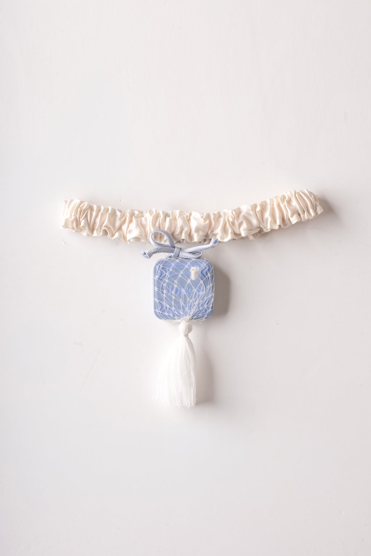 The Jones / Saint Sintra Alpine Garter is a cream-colored silk charmeuse garter featuring a light blue square charm encased in white netting, suspended from a small bow with a white tassel at its base.