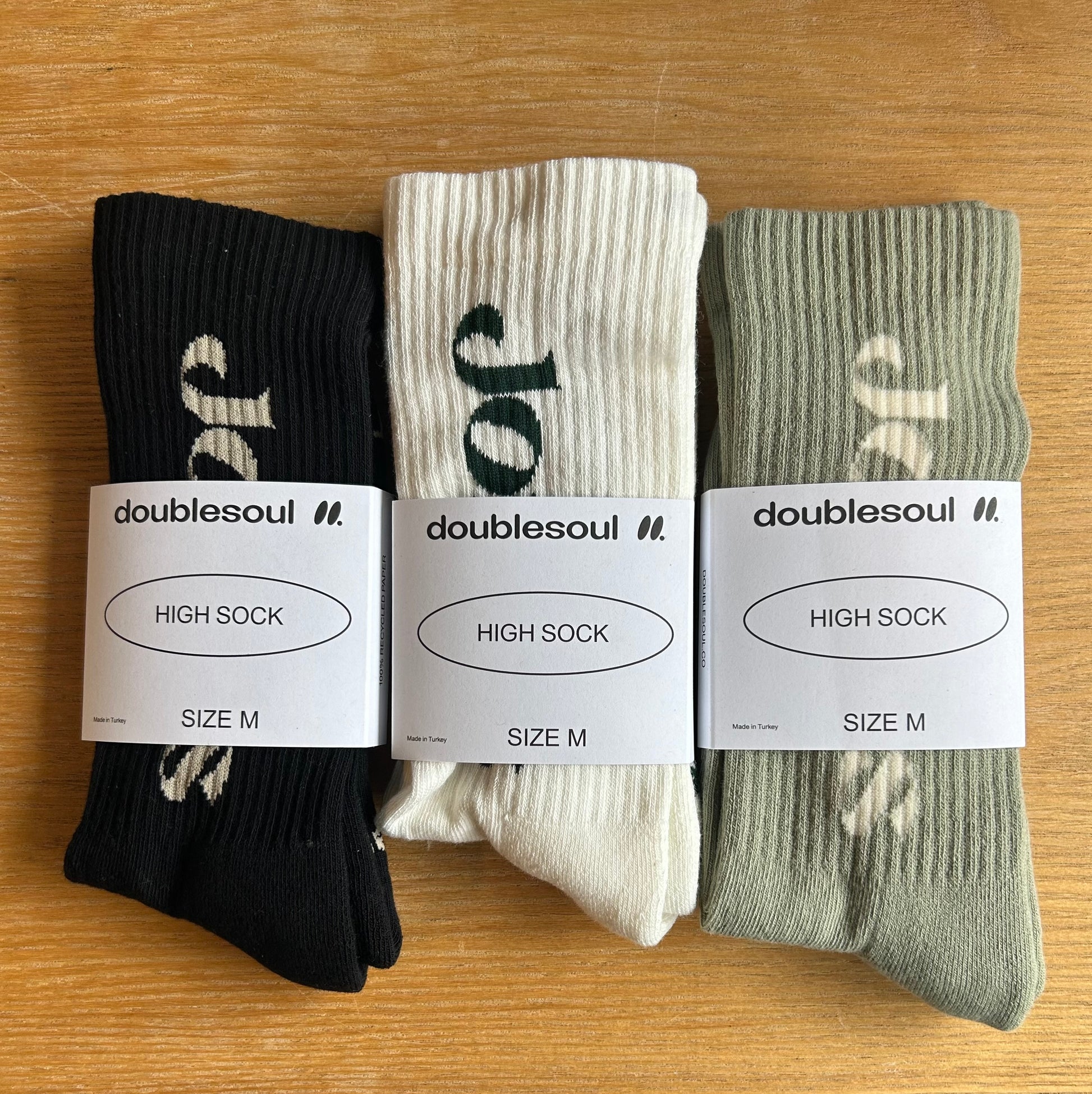 Three pairs of black, white, and green "Merch Jones x Doublesoul Socks" rest on a wooden surface. Tagged as "HIGH SOCK" size M by the brand Jones, these eco-friendly socks might be the comfiest you ever wear.